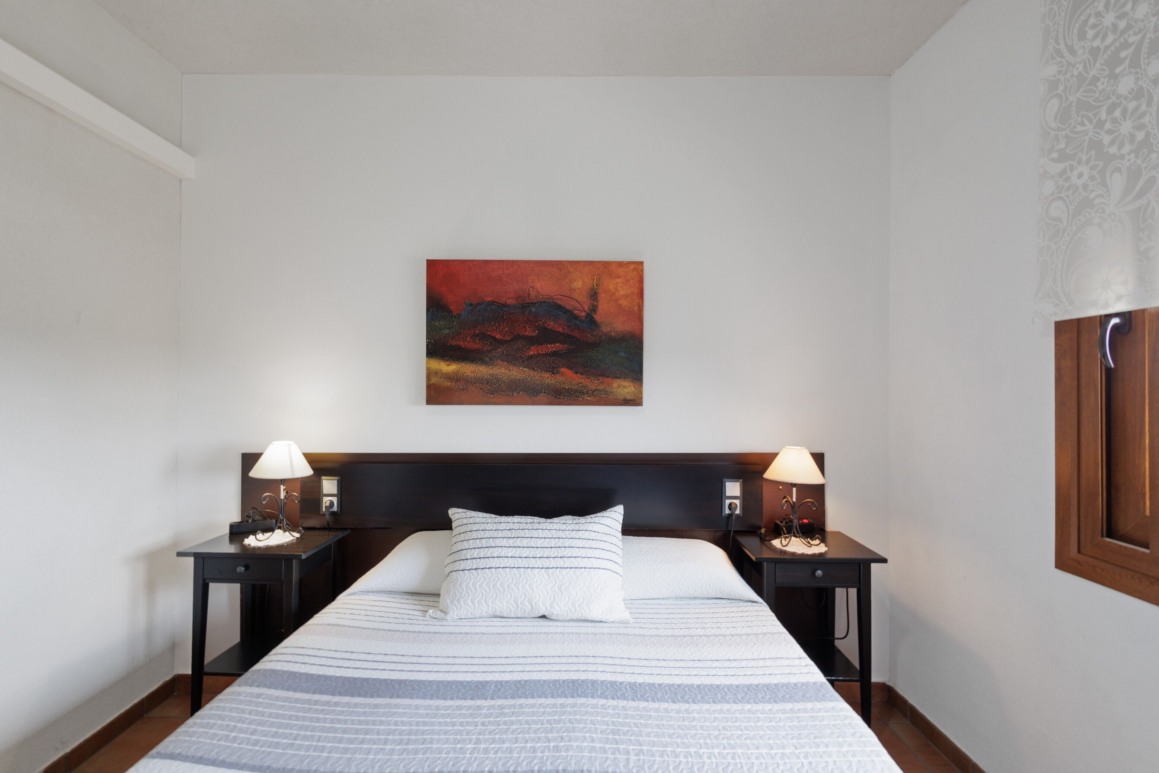 A hotel room with a bed, two bedside tables with lamps, a wall-mounted painting, and a window with a wooden frame.