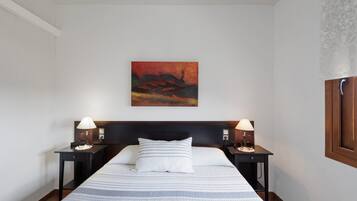 A hotel room with a bed, two bedside tables with lamps, a wall-mounted painting, and a window with a wooden frame.