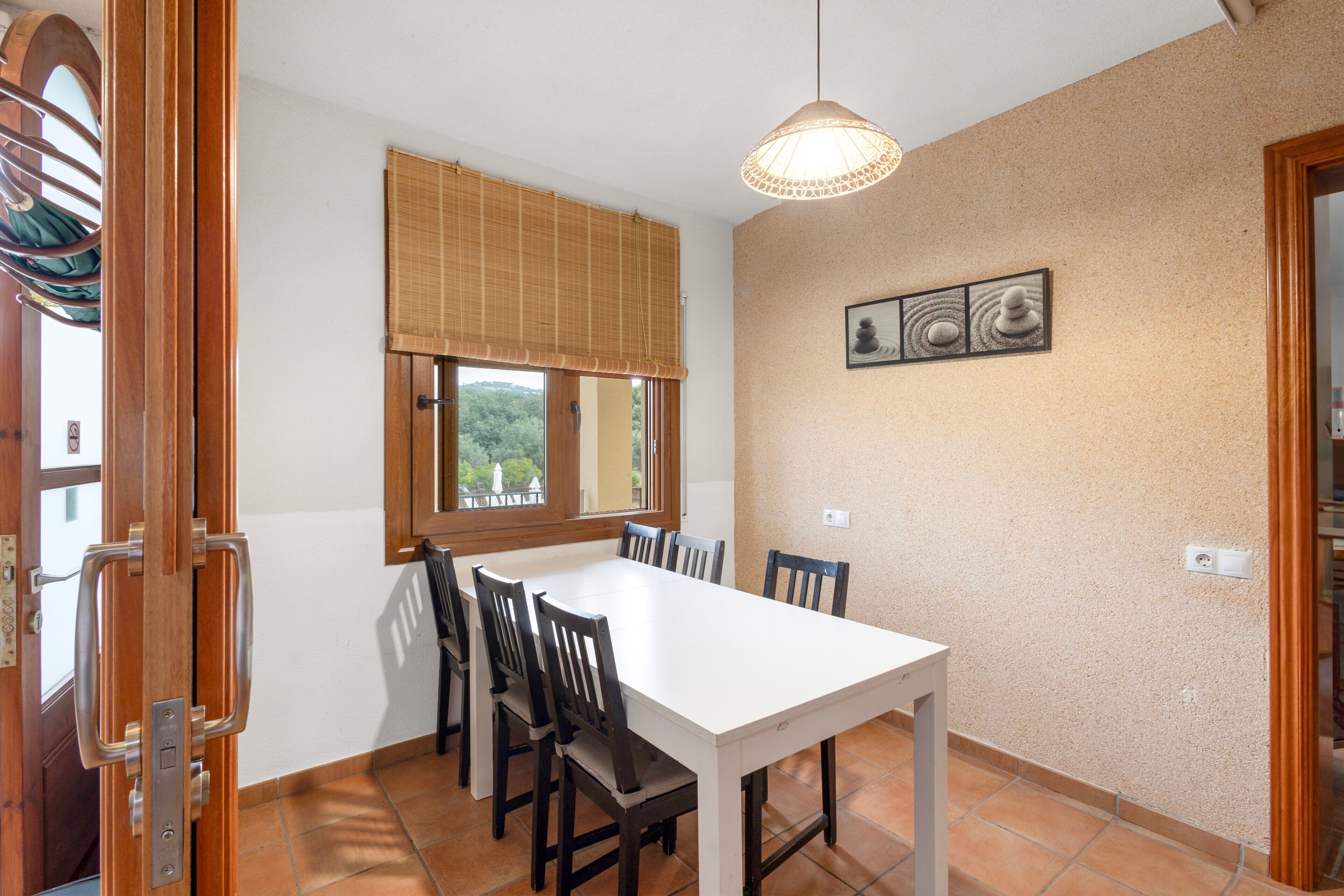Un comedor con una mesa y sillas blancas, una ventana con persianas, una lámpara colgante y un cuadro en la pared.