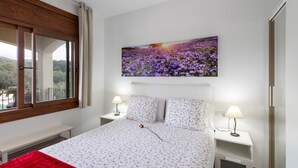 Ein Schlafzimmer mit einem Bett, zwei Nachttischen mit Lampen, einem Fenster mit Vorhängen und einem Wandbild.