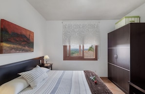 A bedroom with a bed, a nightstand, a wardrobe, a window with a view, and a painting on the wall.