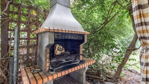 An outdoor brick oven grill with a chimney, surrounded by greenery.