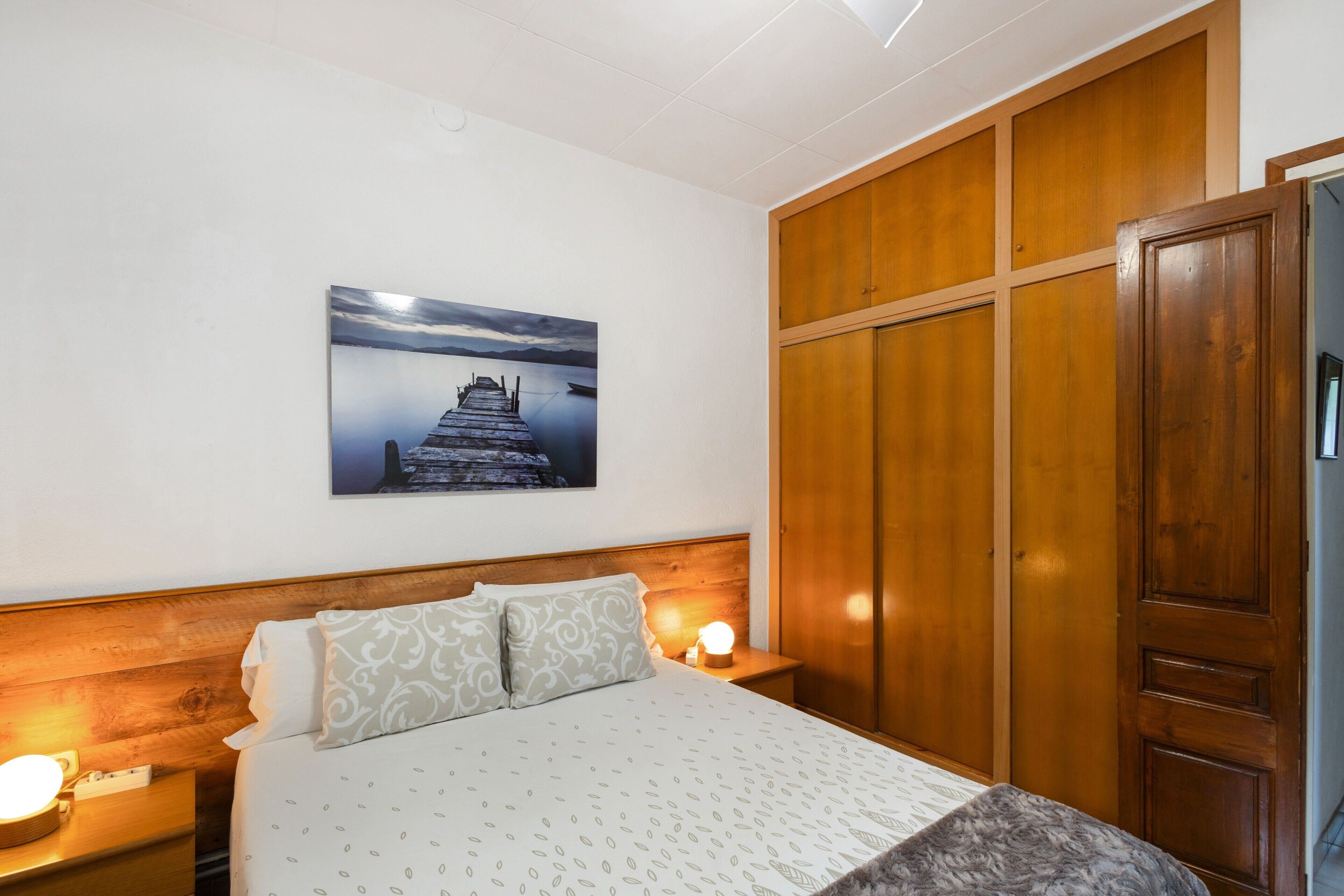 Habitación de hotel con una cama, cabecera de madera, mesita de noche con lámpara, armario y un cuadro en la pared.