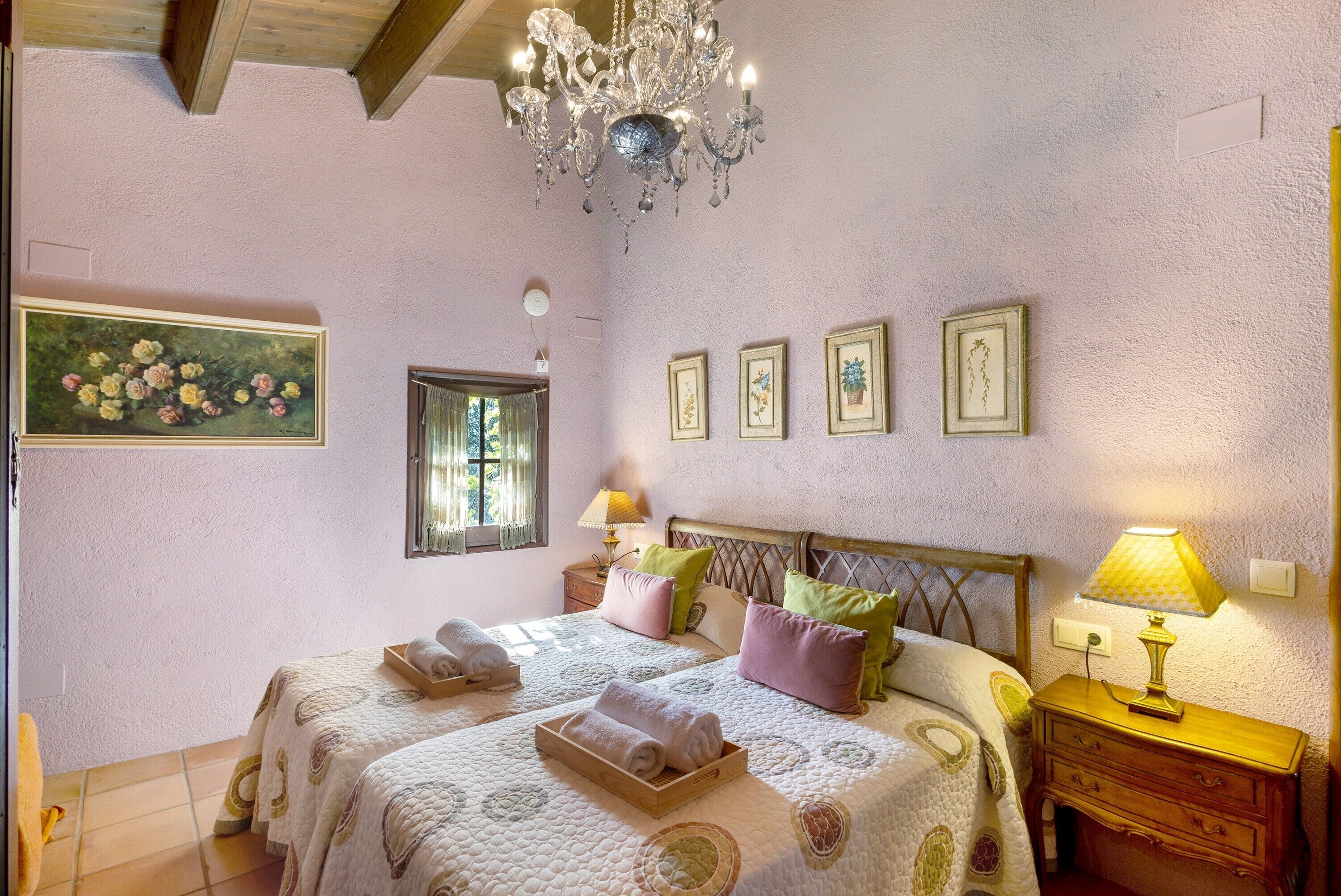 A bedroom with two beds, a chandelier, a window, and wall-mounted artwork.