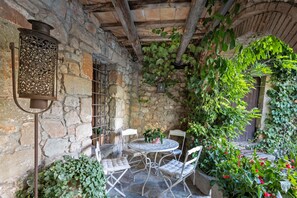 Un patio al aire libre con paredes de piedra, una mesa redonda y cuatro sillas, rodeado de abundante vegetación y un farol de metal.
