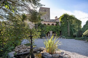 Une cour avec une fontaine, un mur de pierre et un bâtiment doté de fenêtres en arc de cercle et recouvert de lierre.