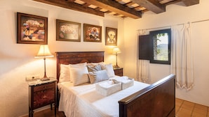 A bedroom with a wooden bed, two bedside tables with lamps, a window with curtains, and three framed artworks on the wall.