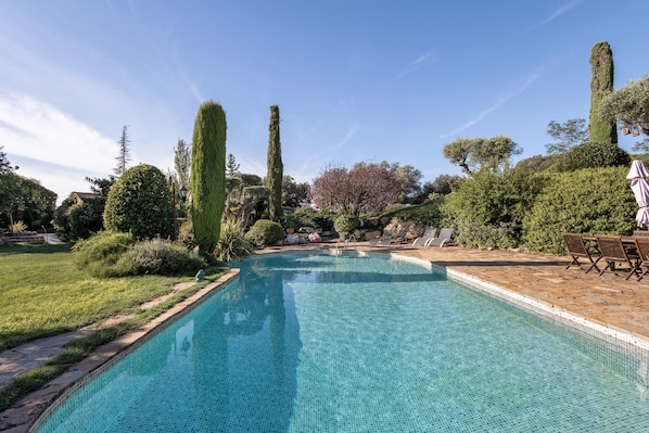 Una piscina con borde de azulejos, rodeada de jardines cuidados y un patio con mobiliario exterior.