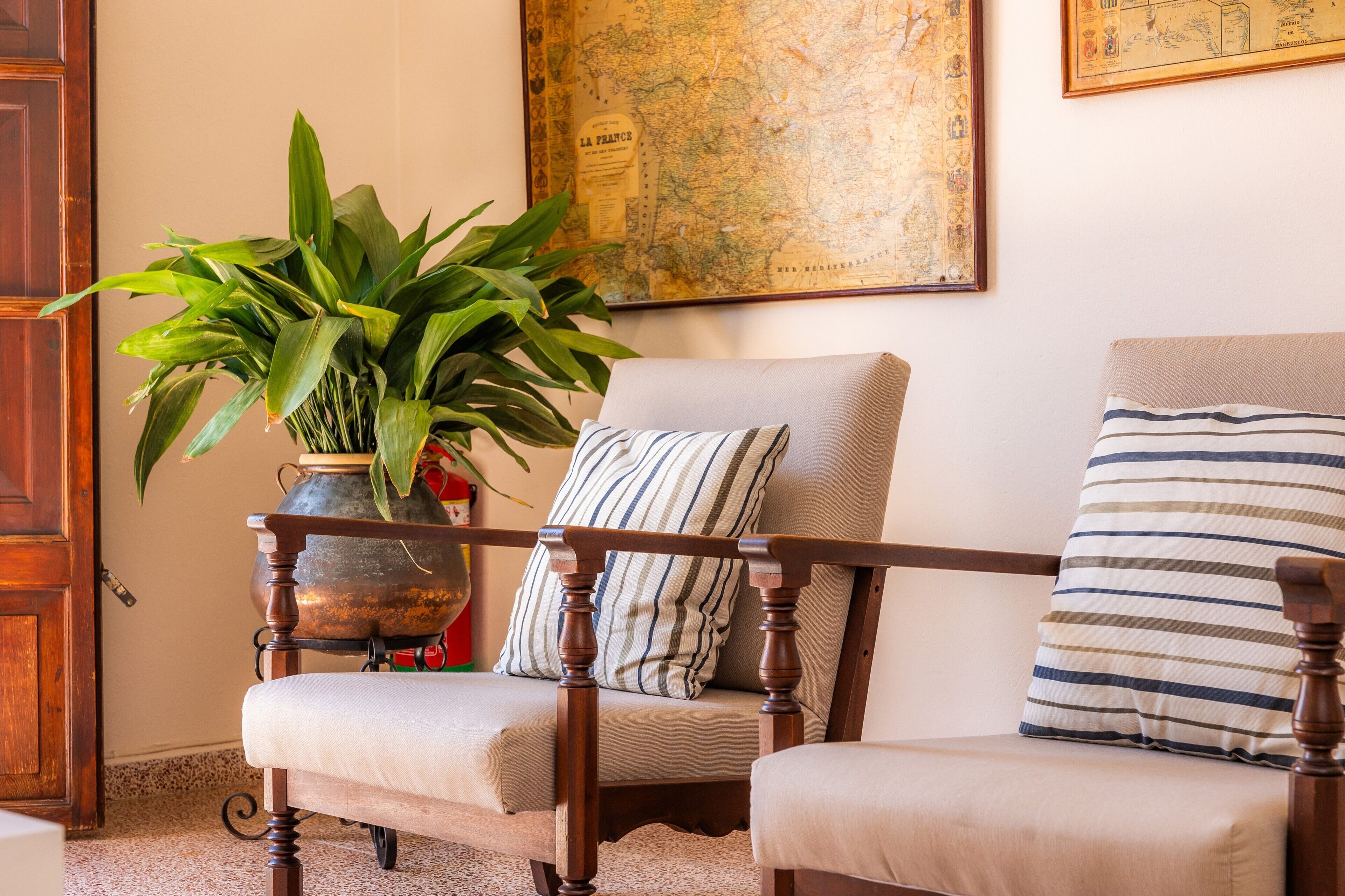 A room with a wooden chair, a cushioned bench, a large potted plant, and a map on the wall.