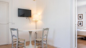 Een kamer met een witte wand, een flatscreen televisie boven een rond tafel met twee stoelen, een vloerlamp en een bed met een witte spreidek.