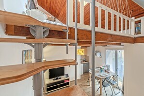 A loft-style room with a wooden ceiling, bunk beds, a television, a dining area, and a kitchen.
