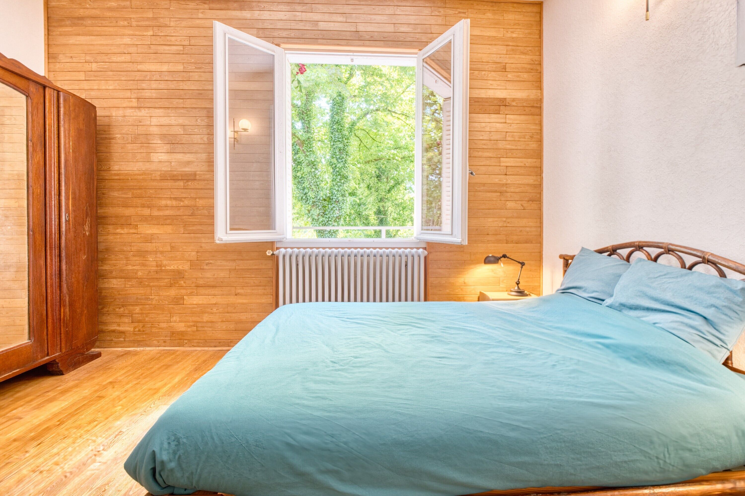 Un dormitorio con piso de madera, una cama con colcha azul, una ventana con persiana abierta y un radiador debajo de la ventana.
