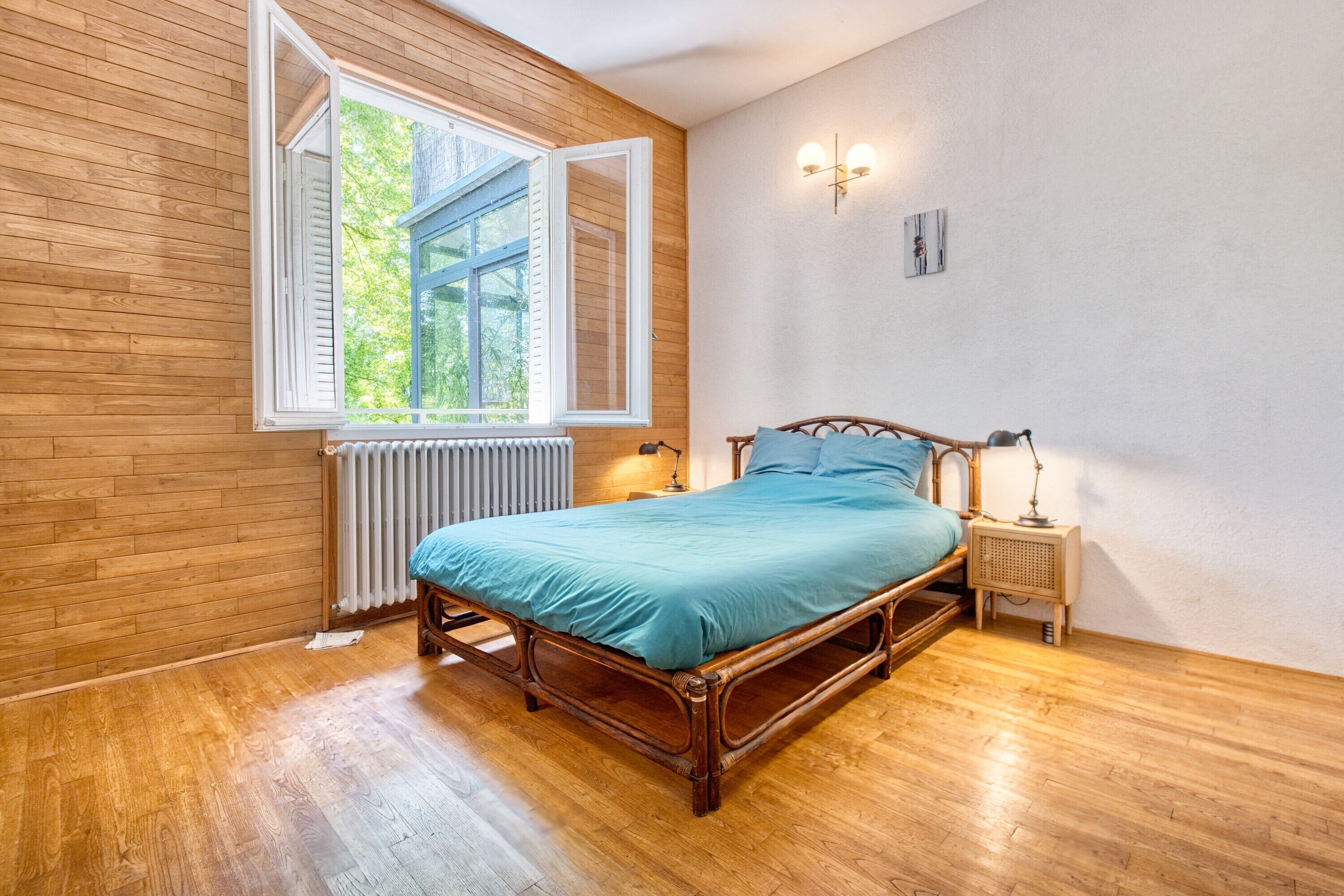 A bedroom with a wooden floor, a bed with a blue bedspread, a window with white shutters, a radiator, and a bedside table with a lamp.