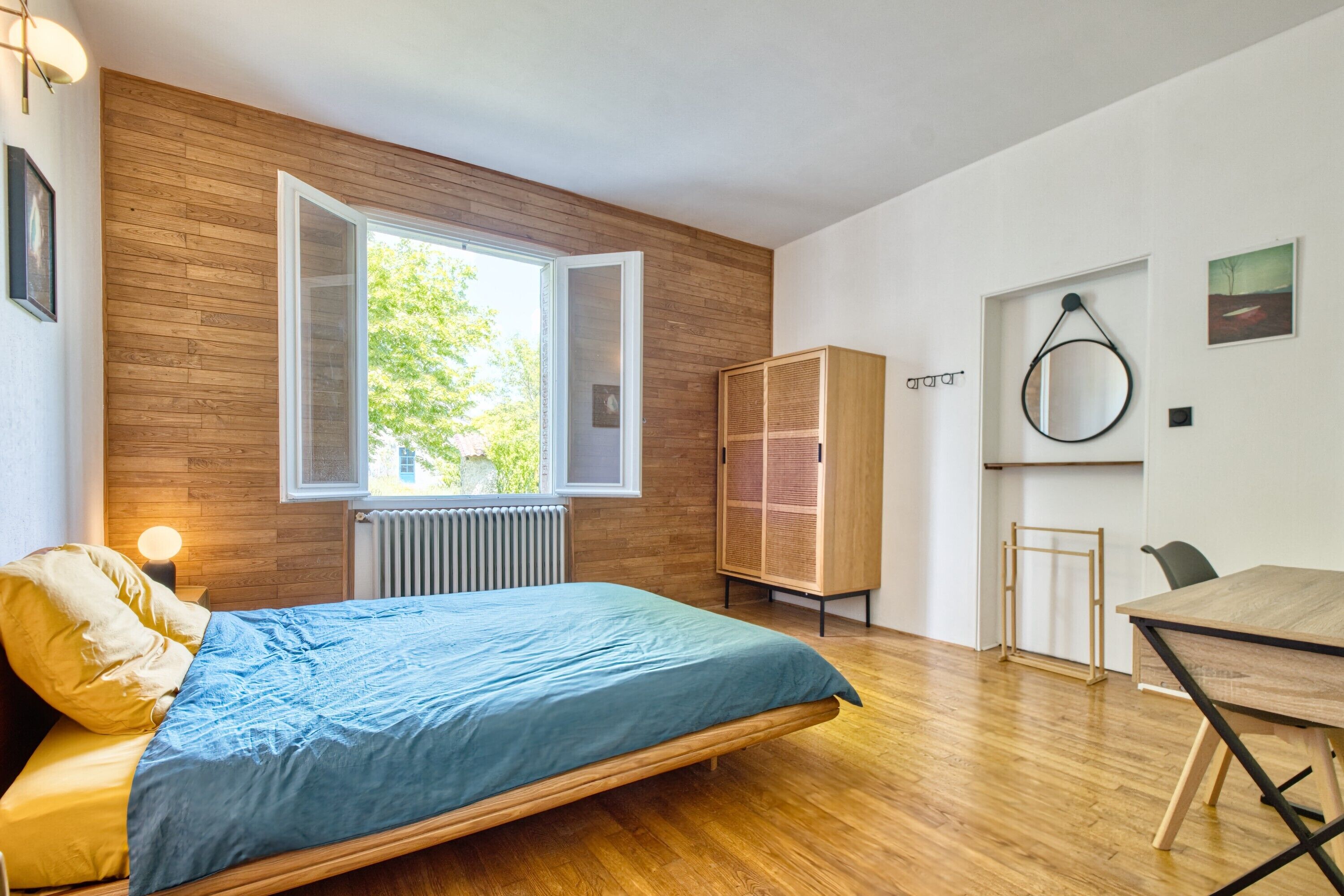 A modern bedroom with a wooden floor, a bed with a blue bedspread, a wooden wardrobe, a dining table, and a window with a view of greenery.