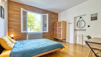 A modern bedroom with a wooden floor, a bed with a blue bedspread, a wooden wardrobe, a dining table, and a window with a view of greenery.