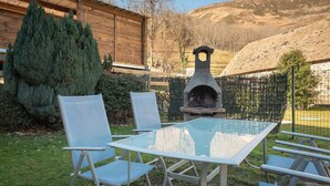 Un patio con una mesa y sillas de vidrio, un horno de piedra, una cabaña de madera y montañas al fondo.