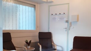 Une pièce comprenant deux fauteuils, une petite table avec une plante, une porte sur laquelle est affichée une note, et une fenêtre avec des stores.