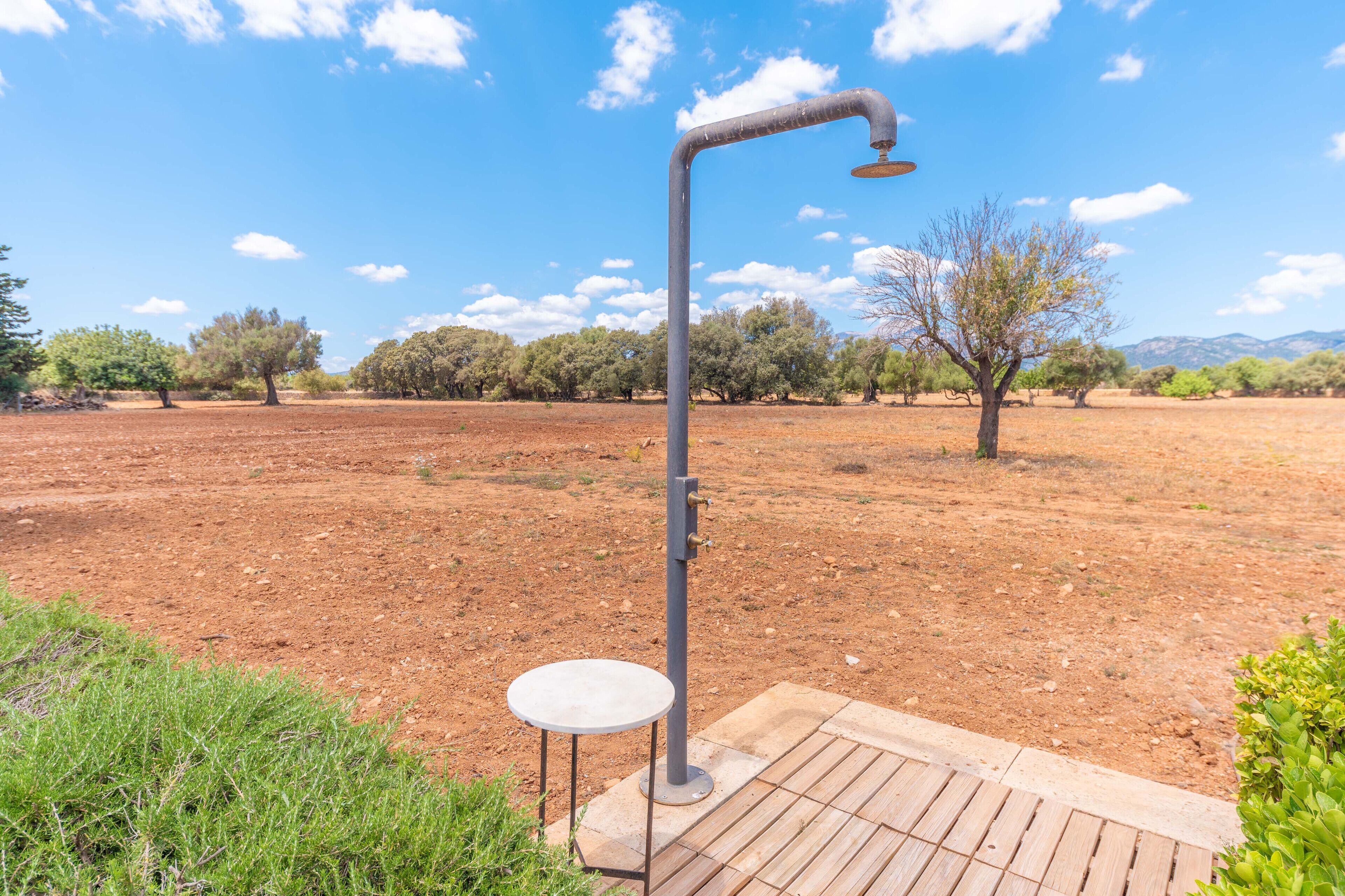 Une douche extérieure en métal, avec un petit tabouret, installée dans un champ aride et dégagé, parsemé de quelques arbres et sous un ciel dégagé.