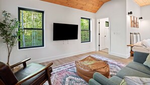A modern living room with a flat-screen TV mounted on the wall, large windows with a view of trees, a wooden ceiling, and a cozy seating area with a patterned rug.