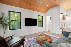 Un salon moderne doté d’un téléviseur à écran plat fixé au mur, de grandes fenêtres offrant une vue sur les arbres, d’un plafond en bois et d’un coin salon confortable agrémenté d’un tapis à motifs.
