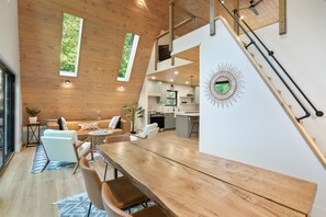 A modern interior with a wooden ceiling, a dining area with a long table, and a kitchen with white cabinets. - The Redhawk | Hocking Hills, Sauna, EV Charger (Logan)