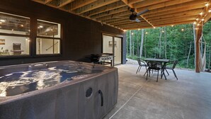 A hot tub in a covered outdoor area with a table and chairs, a ceiling fan, and a view of a forest.