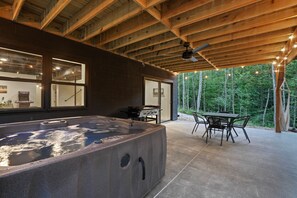 Un bain à remous dans un espace extérieur abrité, équipé d’une table et de chaises, d’un ventilateur de plafond et offrant une vue sur une forêt.