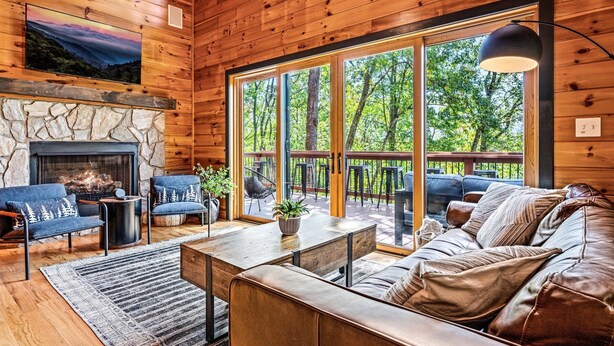 A cozy cabin interior with a stone fireplace, wooden walls, and large windows offering a view of the forest.