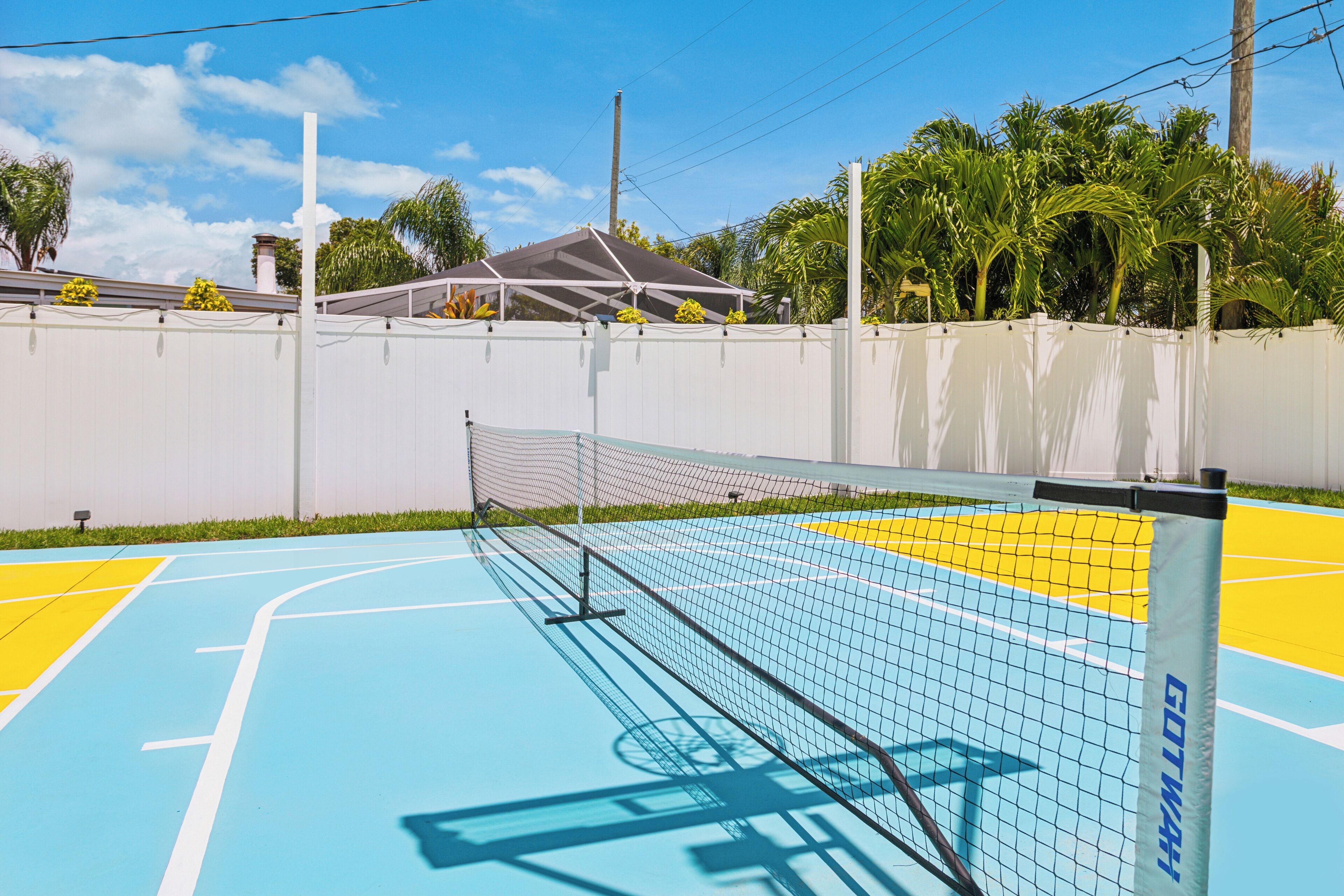 An outdoor tennis court with a white fence, a net, and a blue and yellow playing surface.