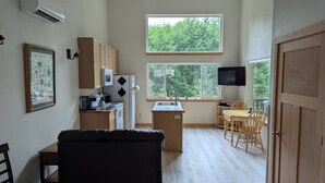 Cocina compacta equipada con microondas, refrigerador y lavamanos. Cuenta con comedor con mesa y sillas, y un amplio ventanal con vista a árboles.