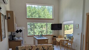 Una habitación amplia con una cama grande, un sofá, una mesa de comedor y un televisor. Hay ventanas que ofrecen luz natural y vistas a la vegetación exterior.