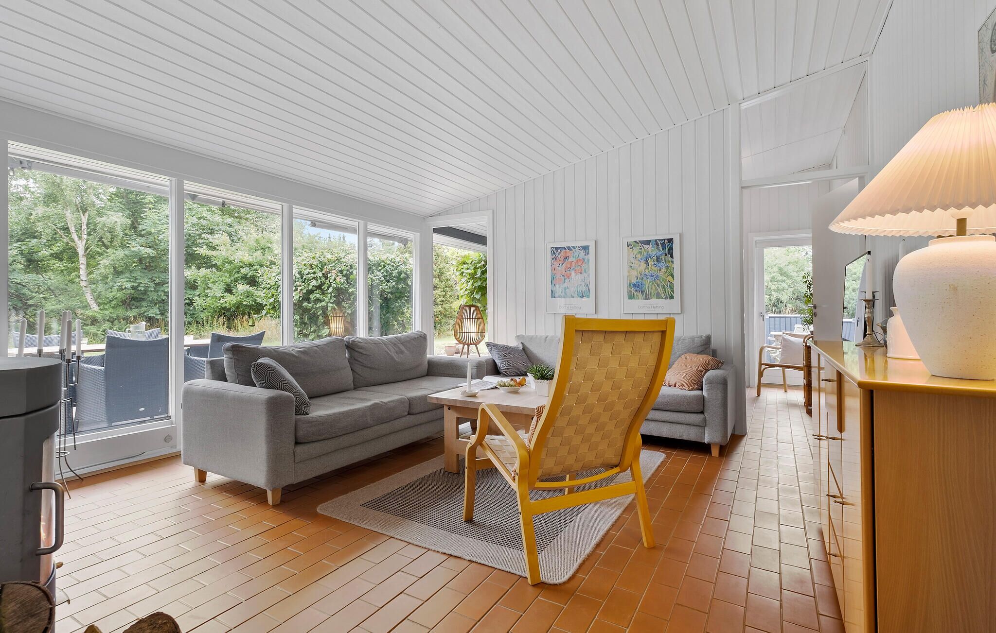Ett modernt vardagsrum med en grå soffa, en gul fåtölj och ett matbord. Det finns stora fönster som ger utsikt över grönområden utanför.