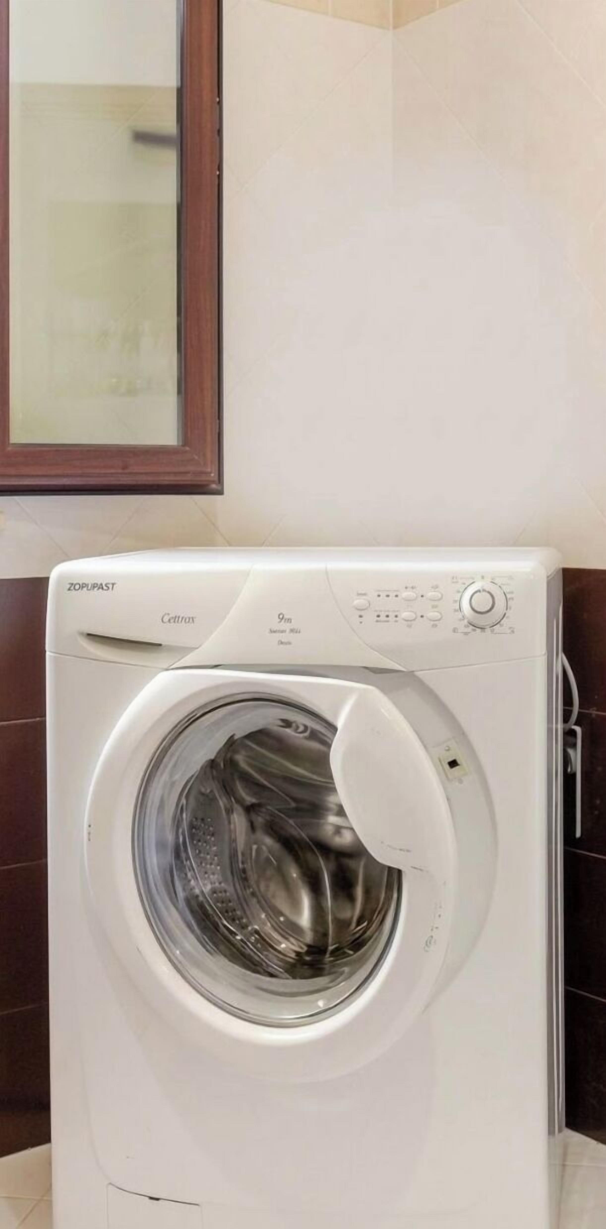 Un lave-linge à chargement frontal doté d’un panneau de commande, situé dans une salle de bain dont les murs sont recouverts de carreaux et qui possède un miroir avec un cadre en bois.