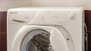 Un lave-linge à chargement frontal doté d’un panneau de commande, situé dans une salle de bain dont les murs sont recouverts de carreaux et qui possède un miroir avec un cadre en bois.