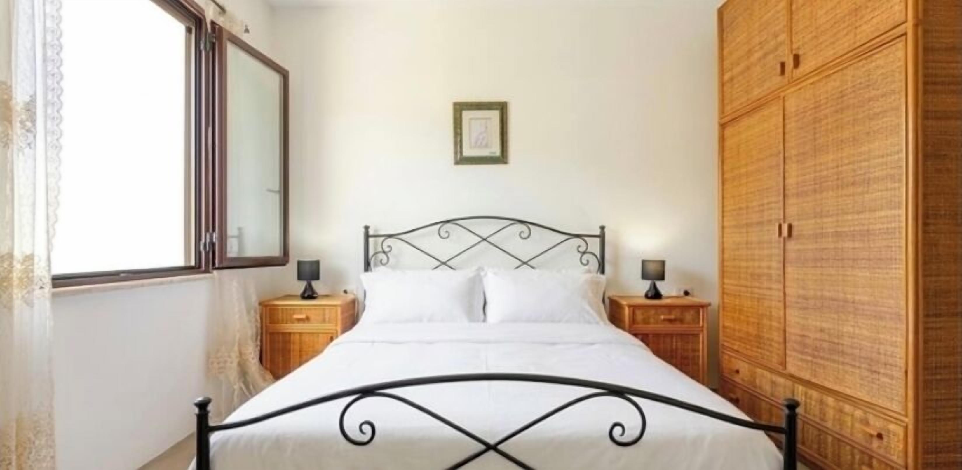 Une chambre à coucher comprenant un lit, deux tables de chevet, une armoire en bois, une fenêtre et un tableau encadré accroché au mur.