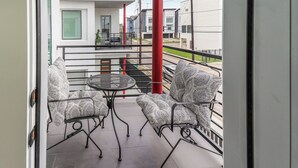 A balcony with two patterned chairs and a small table, overlooking a modern building with a red pillar.