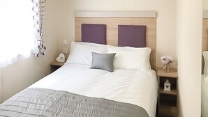 A neatly made bed with a grey quilt, a white pillow, and a grey accent pillow. There is a nightstand with a white lamp and a small decorative item.