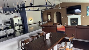 Een moderne keuken met roestvrijstalen apparatuur, een eetgedeelte met een houten tafel en stoelen, een woonkamer met een bank en open haard, en een grote televisie aan de muur.