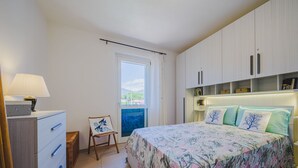 Een slaapkamer met een bed, nachtkastje, stoel, dressoir en een raam met uitzicht.