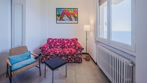 Un salon avec un canapé à motifs floraux rouges, un fauteuil en osier avec des coussins bleus, une petite table noire, un lampadaire et une grande fenêtre donnant sur la mer.