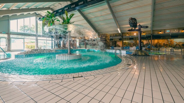 Piscina interior con un elemento central de agua, rodeada por un suelo de baldosas y un techo alto con tragaluces.