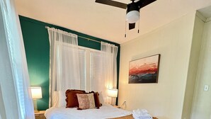 Un dormitorio con cama, ventilador de techo, mesita de noche, lámpara, un cuadro y un tapete estampado.