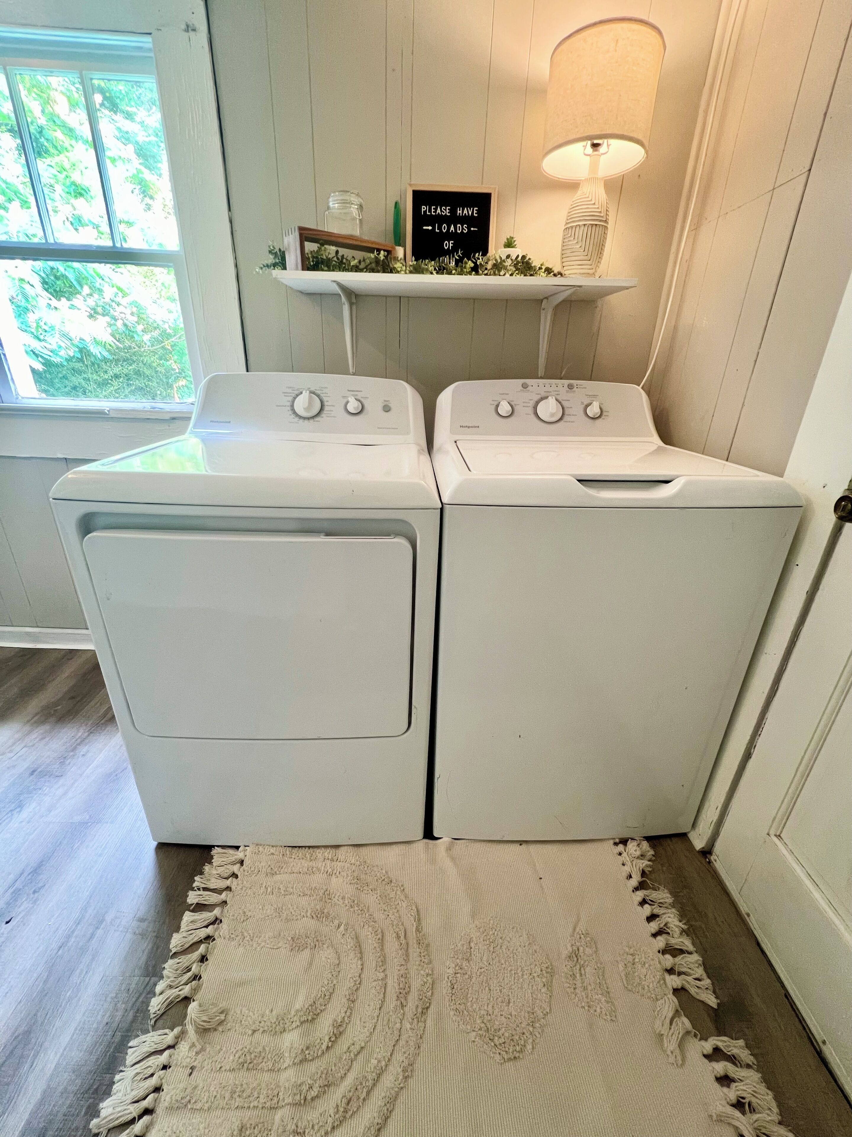 A laundry room with two front-loading washers and dryers, a small shelf with a sign, a lamp, and a rug on the floor.