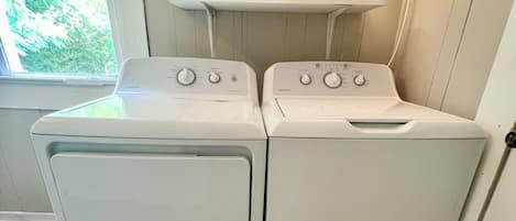A laundry room with two front-loading washers and dryers, a small shelf with a sign, a lamp, and a rug on the floor.