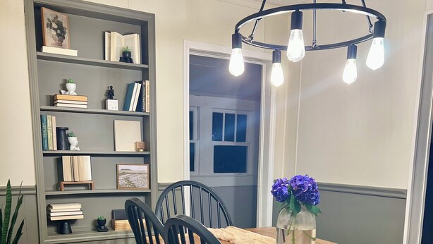 Un comedor con una mesa de madera, sillas negras, una araña y una estantería con libros y objetos decorativos.