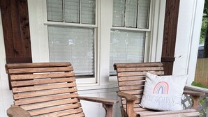 Dos sillas Adirondack de madera, con un cojín en una de ellas, situadas en un porche de pared blanca y ventanas con persianas.