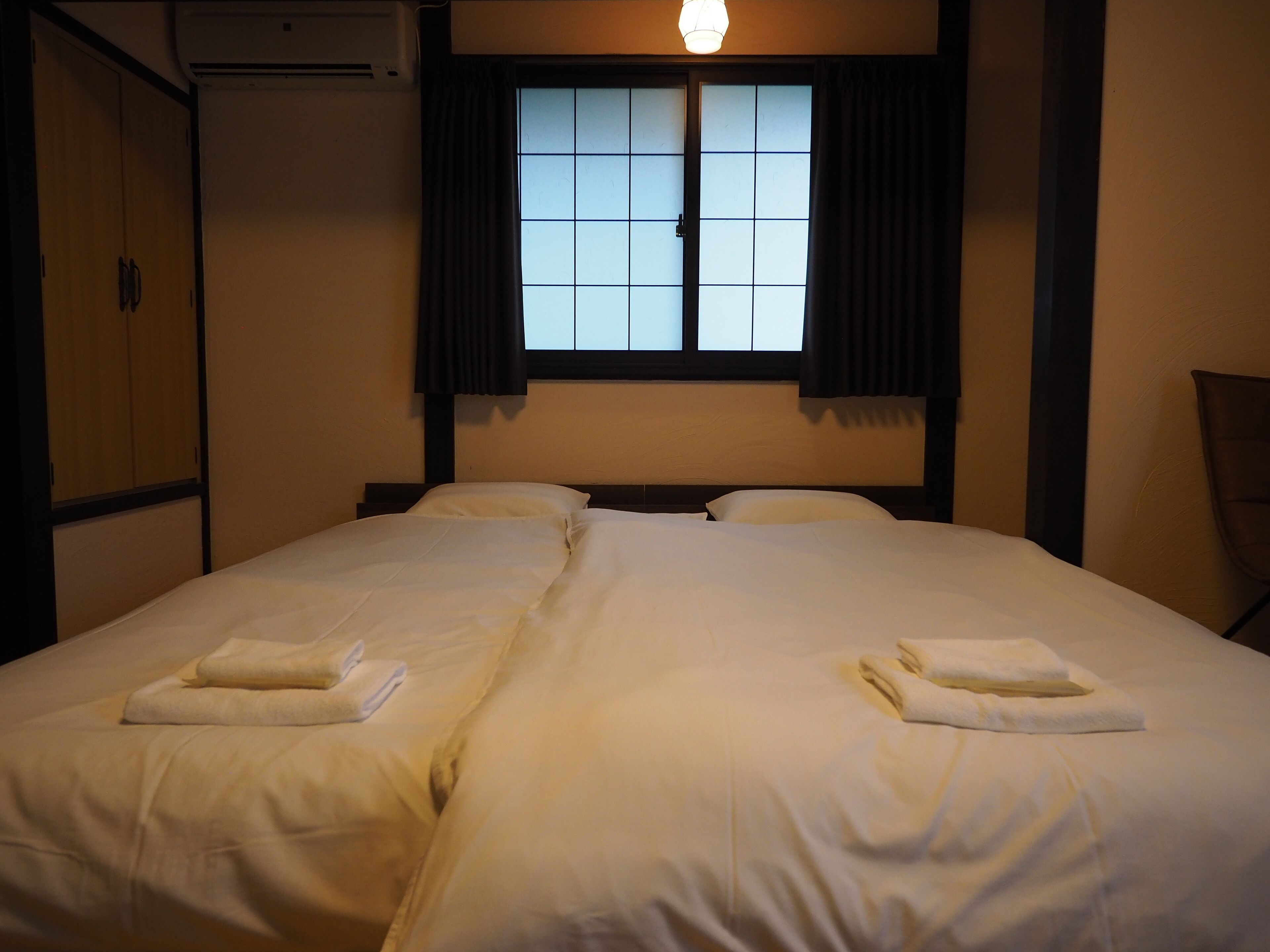 A hotel room with two beds, a window with curtains, a wardrobe, and an air conditioning unit.
