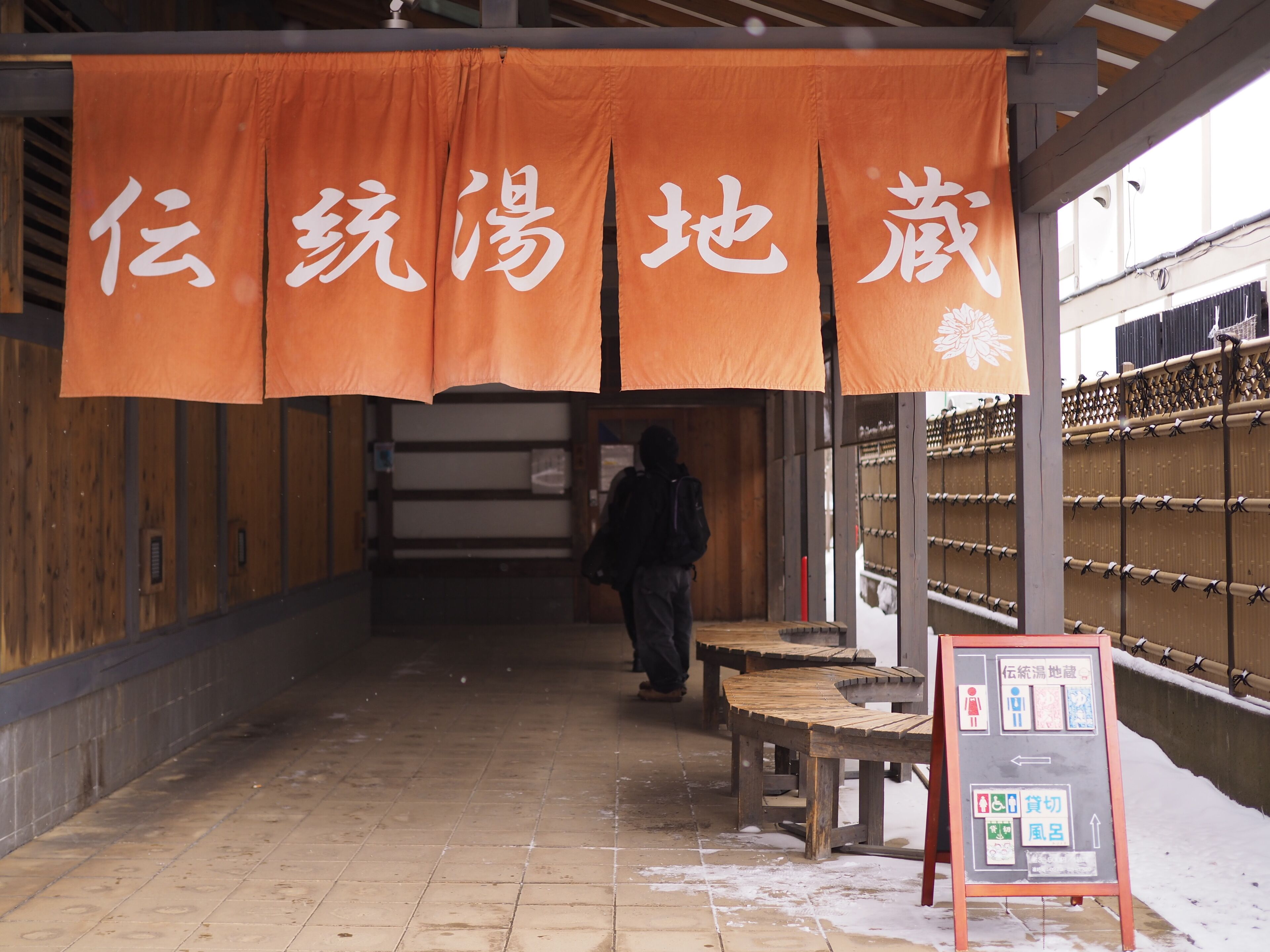 Une entrée abritée, ornée de banderoles orange, de bancs en bois et d’un panneau affichant des symboles.