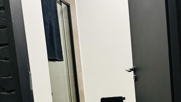 Un couloir étroit avec une porte en verre donnant sur une salle de bain, une serviette noire accrochée au mur et un petit tapis sur le sol.
