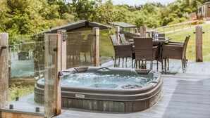 A hot tub on a wooden deck with wicker chairs and a table, surrounded by greenery.